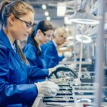 Female Electronics Factory Workers in Blue Work Coat and Protective Glasses Assembling Printed Circuit Boards for Smartphones with Tweezers. High Tech Factory with more Employees in the Background.